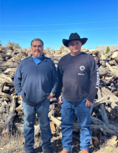 Two men standing in front of large wood pile